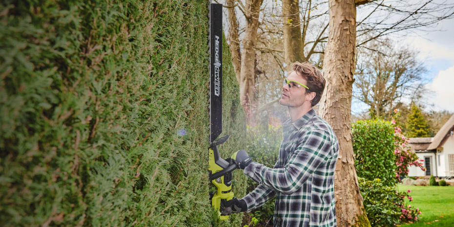 Person wearing safety glasses whilst using a RYOBI hedge trimmer on tall hedge.