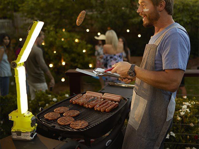 A man stands grilling burgers and sausages on a barbeque, with his Ryobi lamp lighting the space
