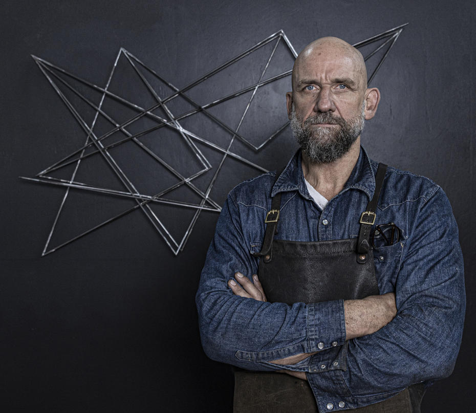 Dion Horstman stands in overalls with arms folded in front of his RYOBI metal artwork
