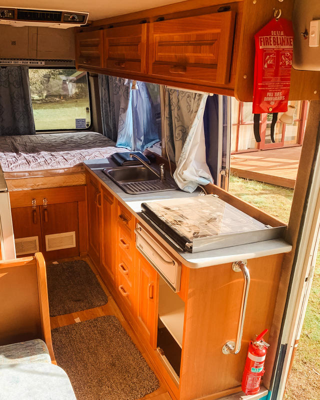 Inside of a campervan showing cooktop, windows, bed and brown dated cupboards
