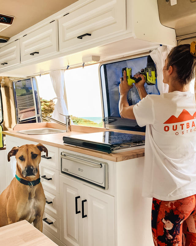 Woman in t-shirt using RYOBI drill on white campervan cupboards. Large dog with her looking towards the camera.