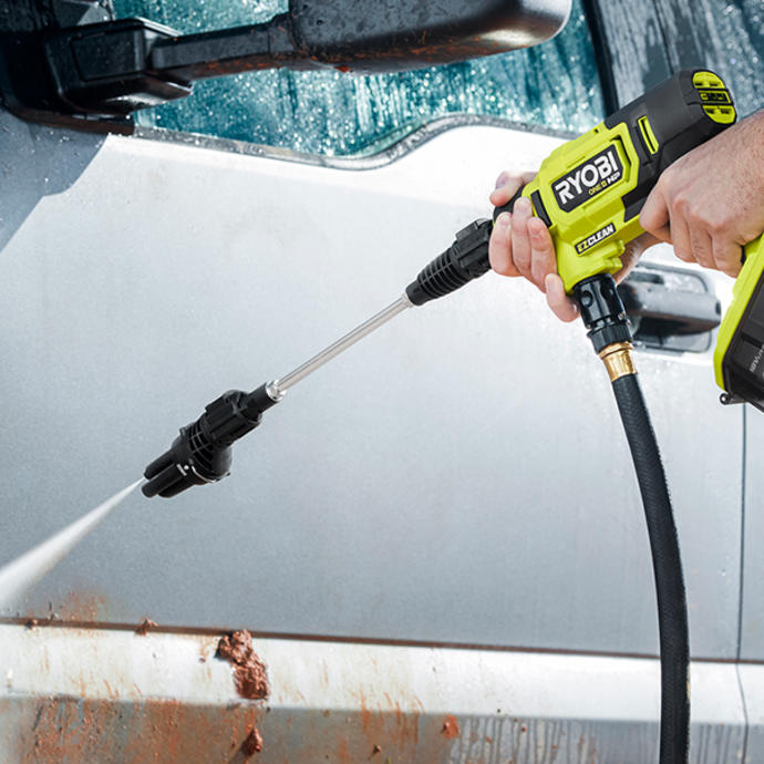 A Ryobi pressure washer being used to clean a dirty, grey car