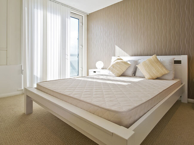 A bedroom with a light, neutral colour scheme with a white timber bedframe and unmade mattress in the centre of the room. 
