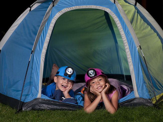 A young boy and girl wearing coloured safety hats with headlamps, laying down in a camping tent on a grassed area. 