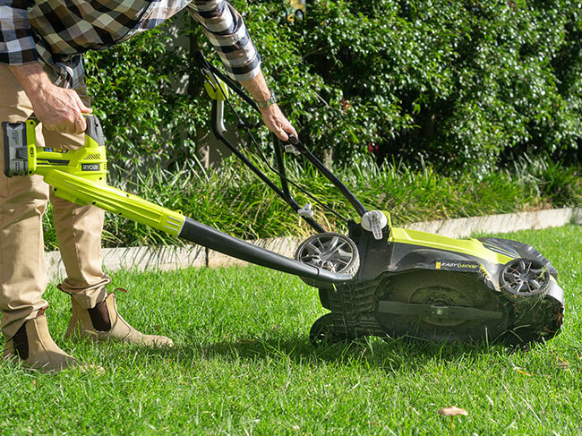 A man using a RYOBI garden blower to clear debris from the underside of a lawn mower. 