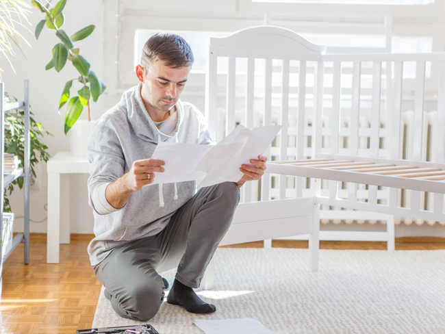 A man kneeling down in a bedroom next to a crib, reading instructions for how to assemble the crib. 