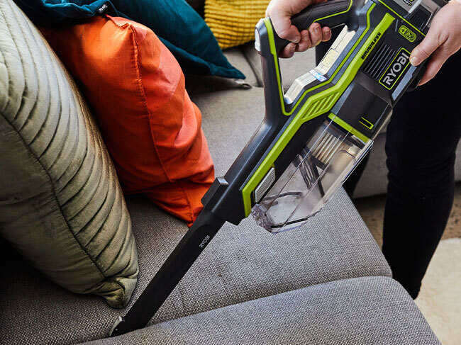 A RYOBI hand vacuum being used with a crevice tool to clean between the seat cushions on a couch. 