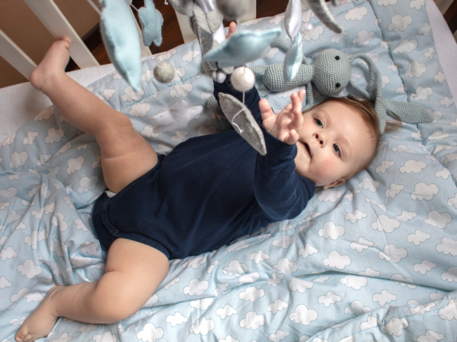 A baby boy in a onsie laying in a crib, reaching up for a toy mobile. A cloud-covered blanket lines the crib. 