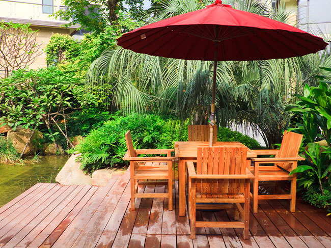 A timber outdoor table setting covered by a red umbrella, located on a backyard timber deck surrounded by greenery. 