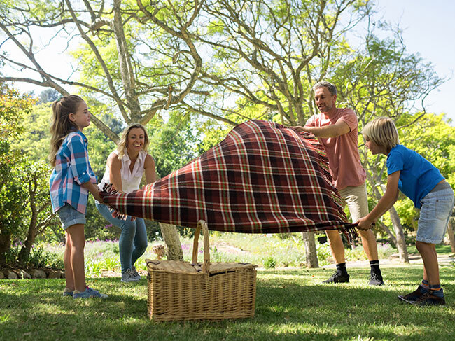 A family of four placing down the corners of a picnic blanket on a grassed area in a park, next to a wicker picnic basket. 