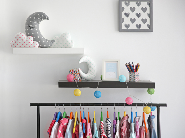 A white wall with a mounted picture frame & floating shelves. Brightly coloured clothes hang on a clothesrack below. 