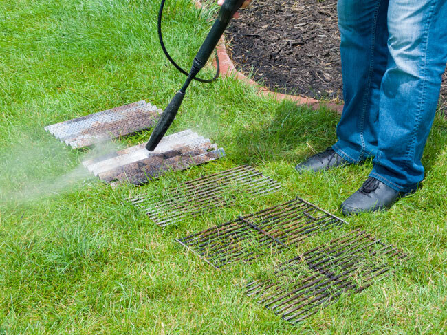 A person standing on a grass lawn using a RYOBI pressure washer to clean barbeque grills and flame diffusers. 