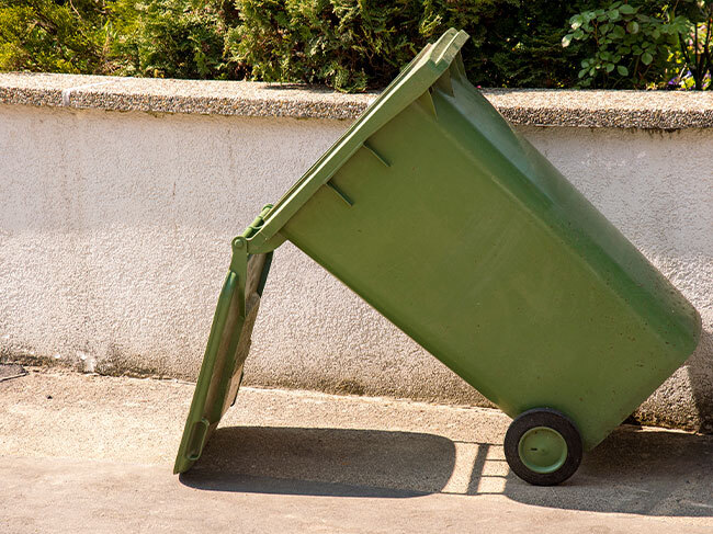 A green wheelie bin in front of a concrete retaining wall, propped at a 45 degree angle by it's open lid. 