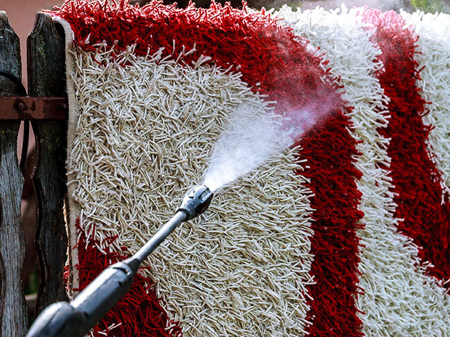 A RYOBI pressure washer being used with a vario lance to clean a floor rug, draped over a timber fence. 