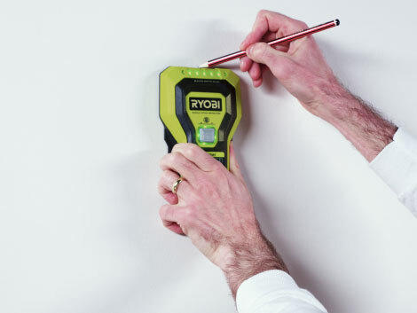 Hands using stud finder and pencil against a white wall.
