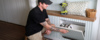 Jake O'Neill opens a draw in the DIY shoe storage cupboard to reveal three pairs of shoes stored neatly. Sitting on the benchtop above the cupboard is a RYOBI drill driver.